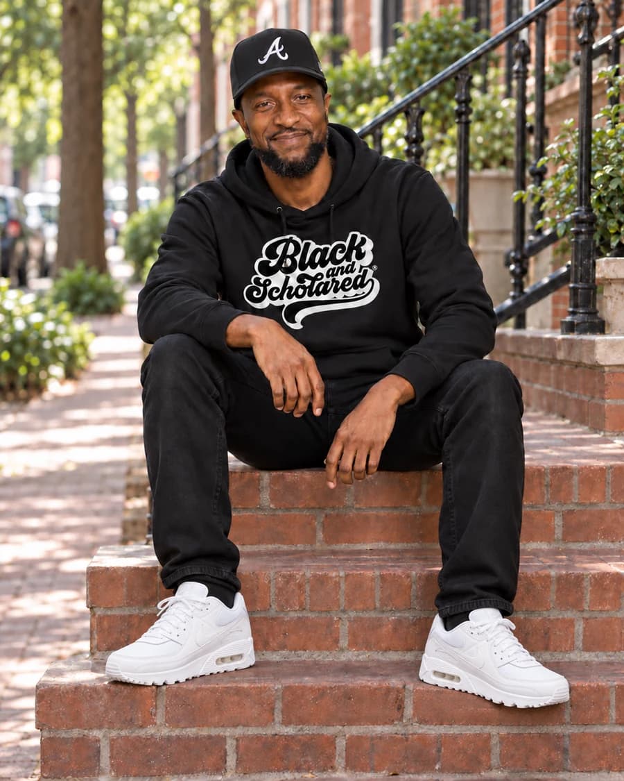 Christopher “CJ” Nurse seated on brick steps in a black Black and Schooled hoodie and cap
